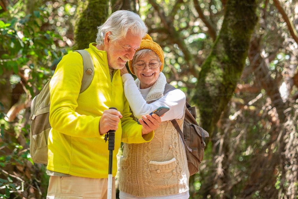 Smiling senior couple hiking in the forest with backpacks looking at mobile phone, elderly couple enjoying healthy lifestyle and retirement