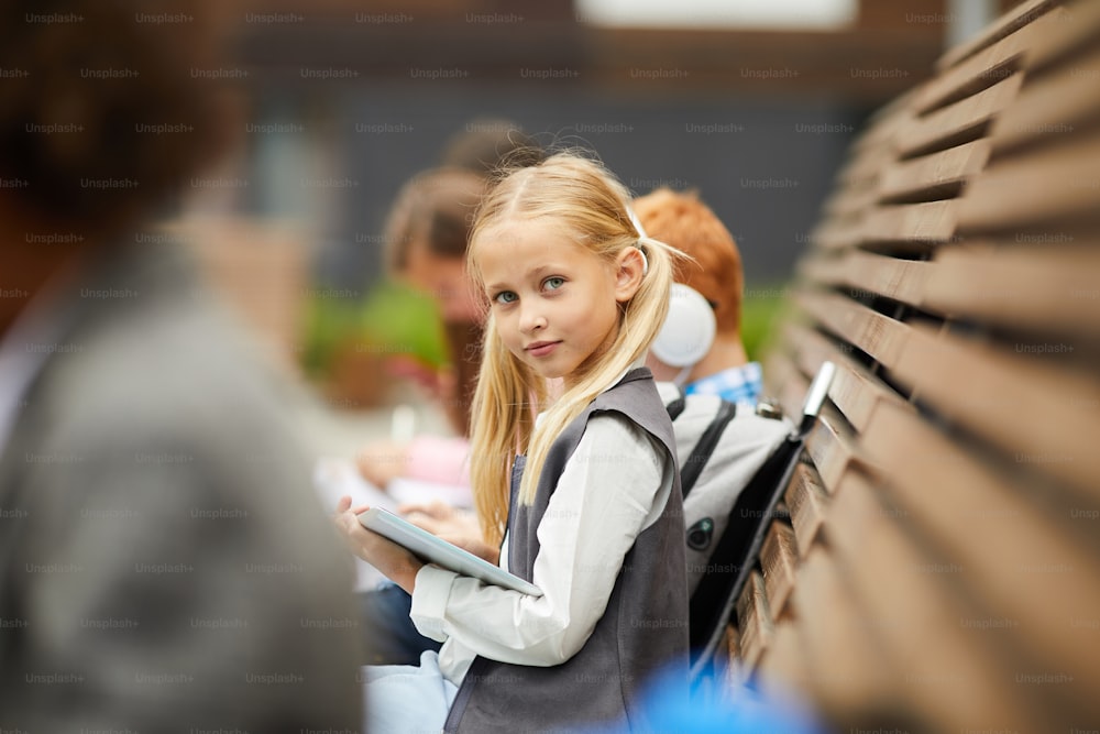 Portrait of schoolgirl with long blond hair looking at camera while sitting on the bench with her classmates and using digital tablet outdoors