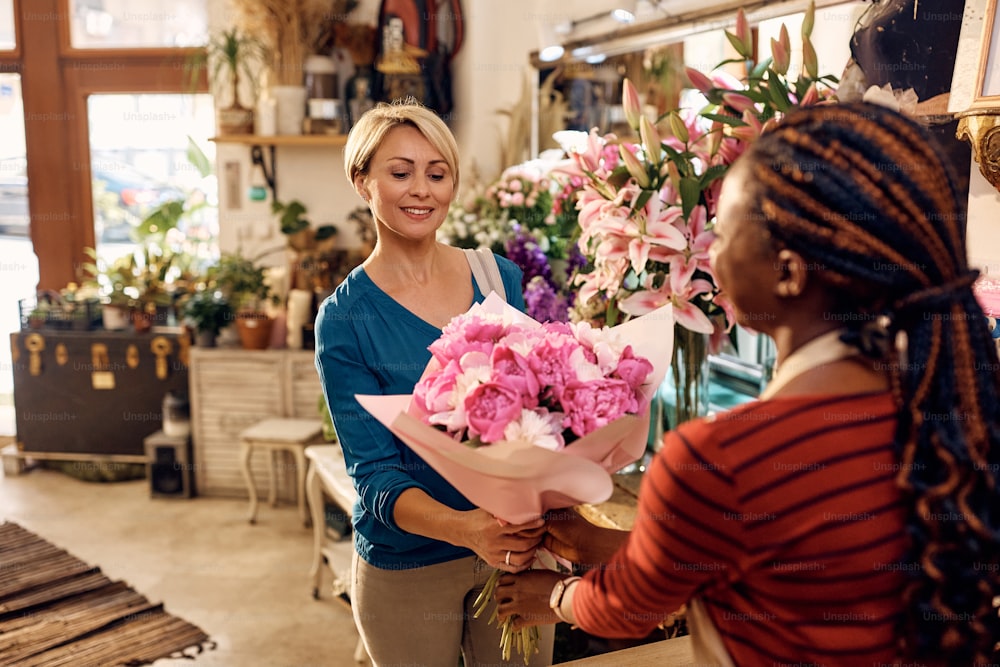 African American florist giving bouquet of fresh flowers to her customer at flower shop. Focus is on female customer.