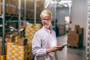 Young Caucasian blonde woman in white uniform using tablet in warehouse.