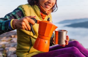 A woman traveler boils a geyser coffee machine and drinks a hot drink from the mug, admiring the colorful dawn with the fog in the mountains, camping and hiking in nature concept