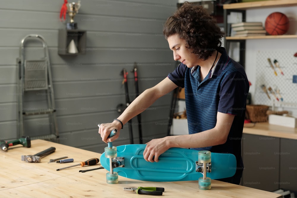 Jugendlicher Typ mit Handwerkzeugen, die Räder auf dem Skateboard montieren, während er am Holztisch in der Garage gegen Trittleiter und andere Haushaltssachen steht