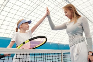 Ragazza adolescente felice in abbigliamento sportivo che tiene la racchetta da tennis mentre dà il cinque al suo allenatore sopra la rete dopo aver giocato con successo sullo stadio moderno