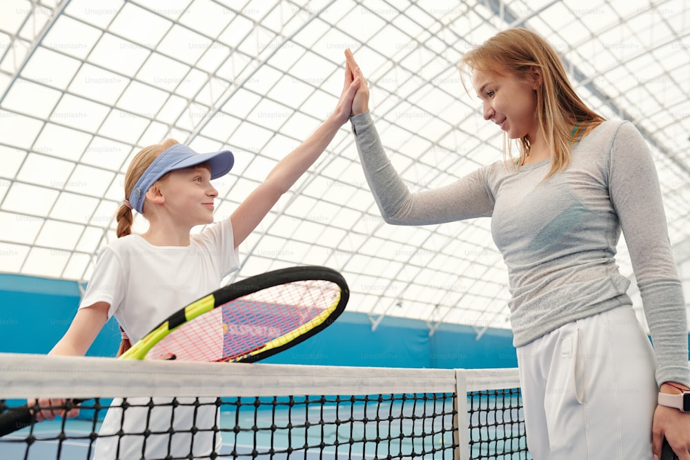 Glückliches Teenager-Mädchen in Activewear, das Tennisschläger hält und ihrem Trainer nach erfolgreichem Spiel im modernen Stadion High Five über das Netz gibt
