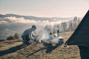 Jeune homme faisant un feu de camp assis près de la tente dans les montagnes