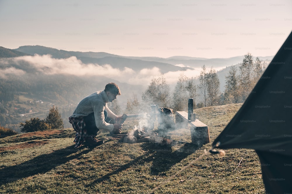 Young man making a campfire while sitting near the tent in the mountains