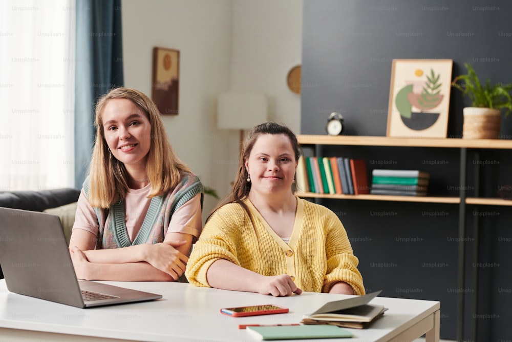 Portrait of young woman with Down syndrome and her female teacher sitting at table with laptop on it at home looking at camera