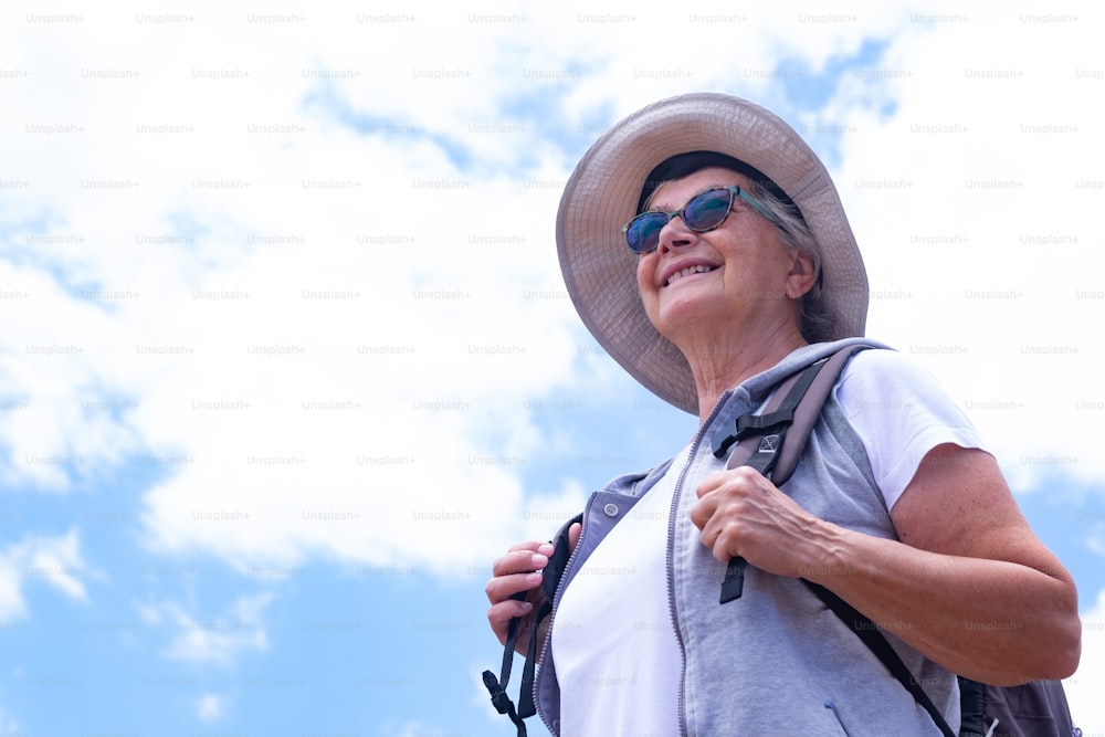 Senior carefree woman with backpack and cap enjoying outdoors excursion. Blue sky