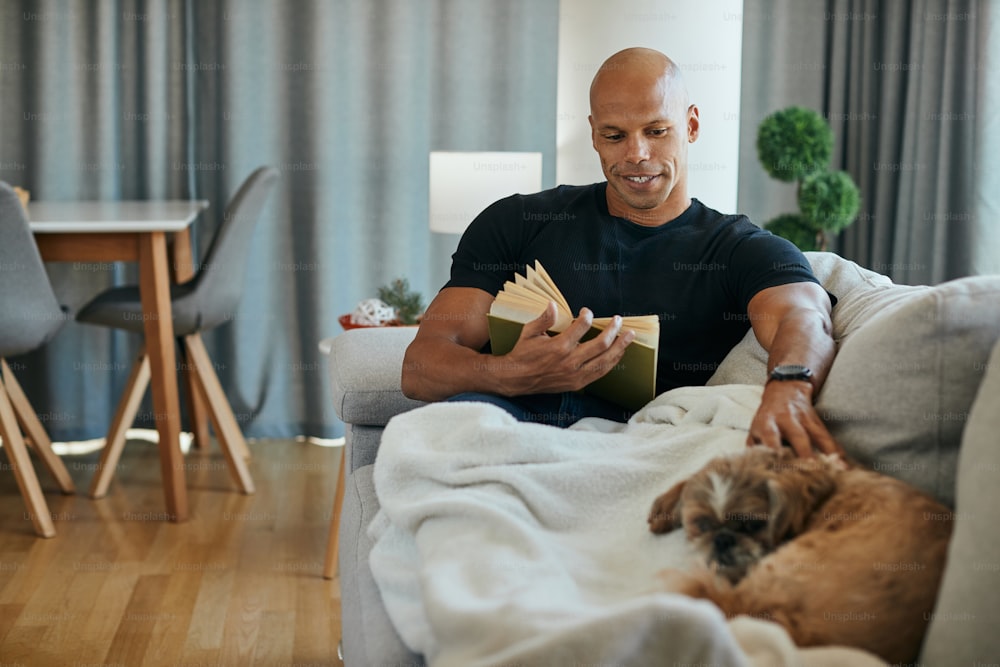 Happy African American man reading book while relaxing with his dog on sofa in the living room.