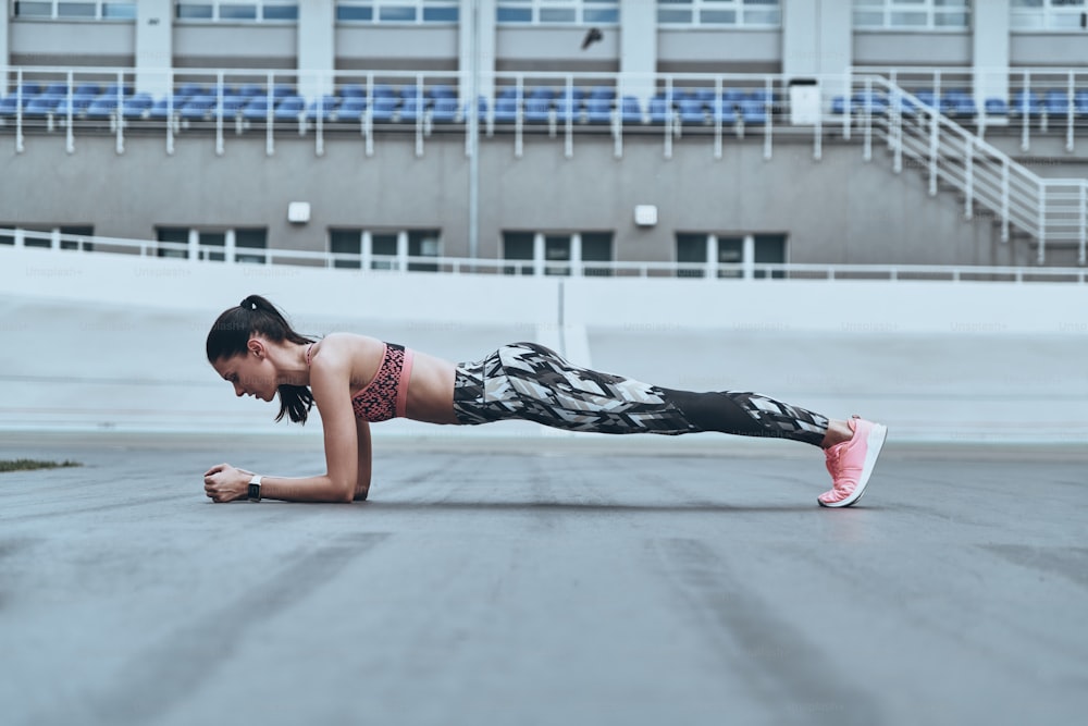 Beautiful young woman in sports clothing keeping plank position while exercising outdoors