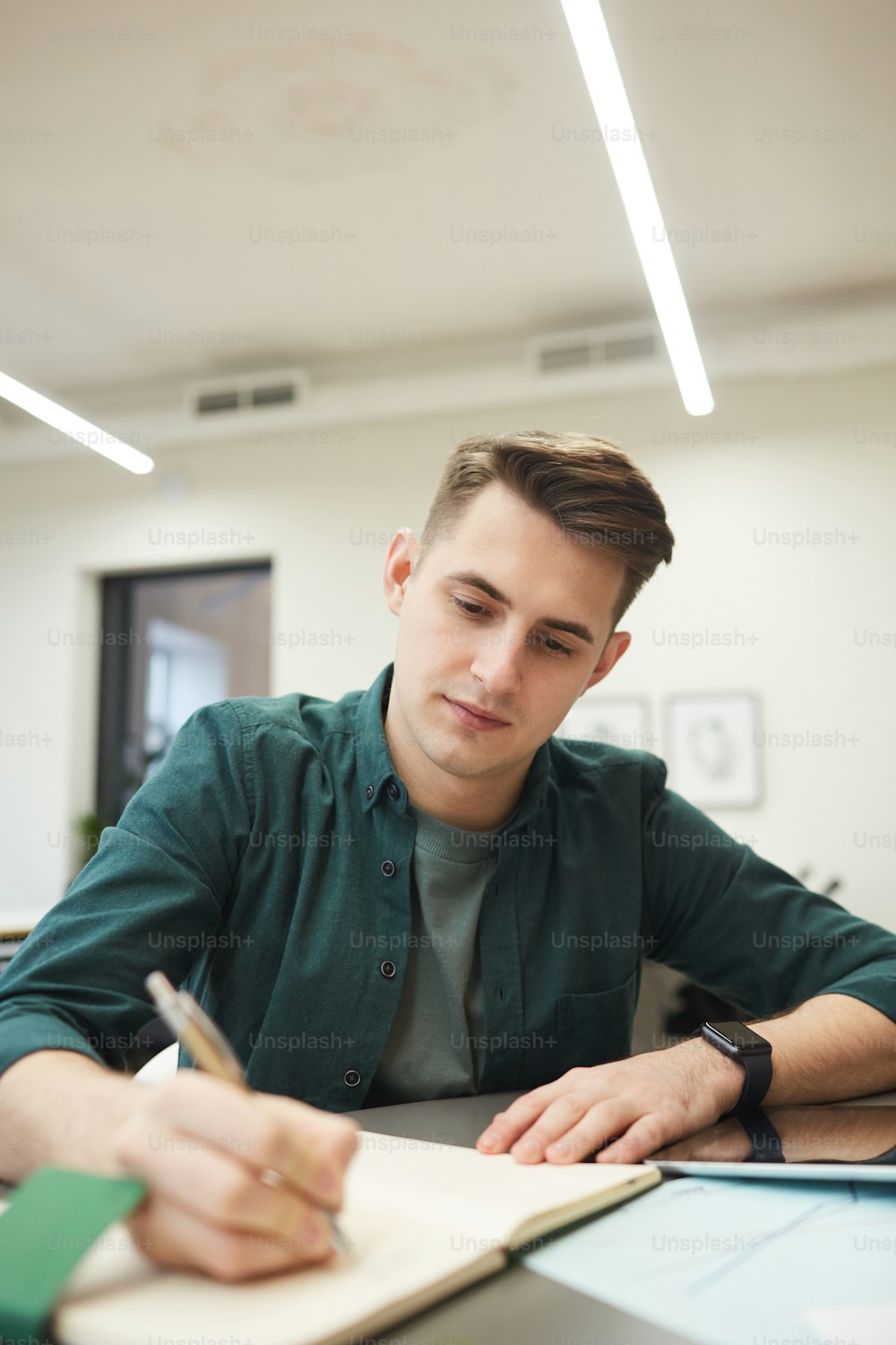 Young man sitting at the table and making notes in notepad while working at office