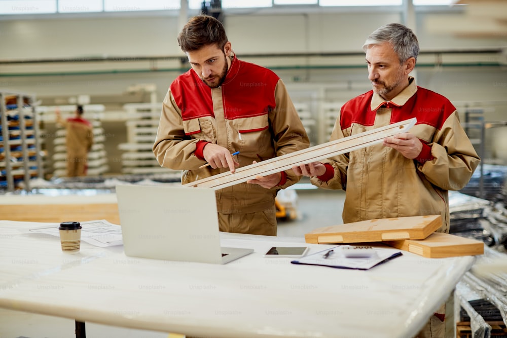 Lavoratori maschi che usano il computer mentre confrontano le misure di un pezzo di legno in un laboratorio di falegnameria.