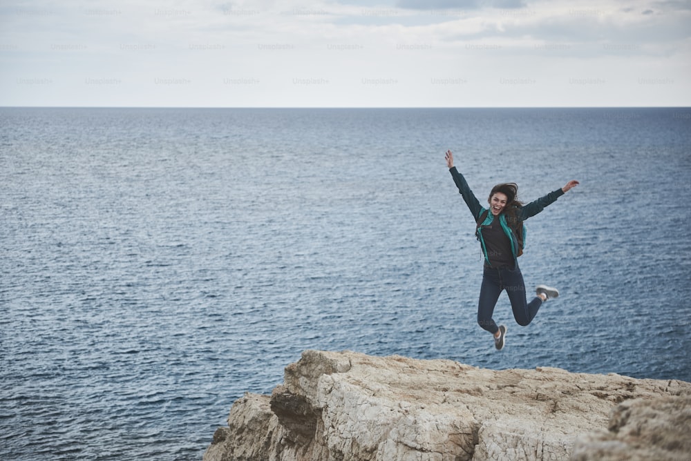 Freedom. Full length portrait of happy girl is jumping on rock mountain with excitement. She is stretching hands up and laughing. Wonderful seascape on background