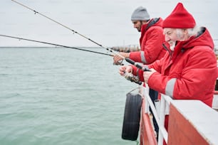 Side view of the happy confident senior man and his young colleague together fishing from a boat at winter weather time in cloudy day under grey sky on the sea. Stock photo
