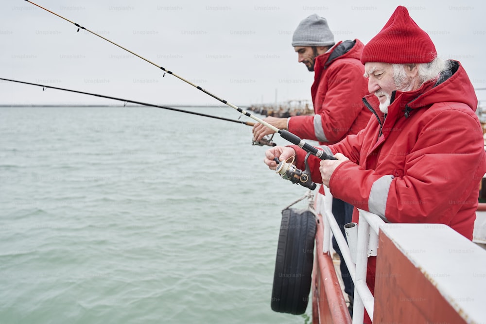 Side view of the happy confident senior man and his young colleague together fishing from a boat at winter weather time in cloudy day under grey sky on the sea. Stock photo