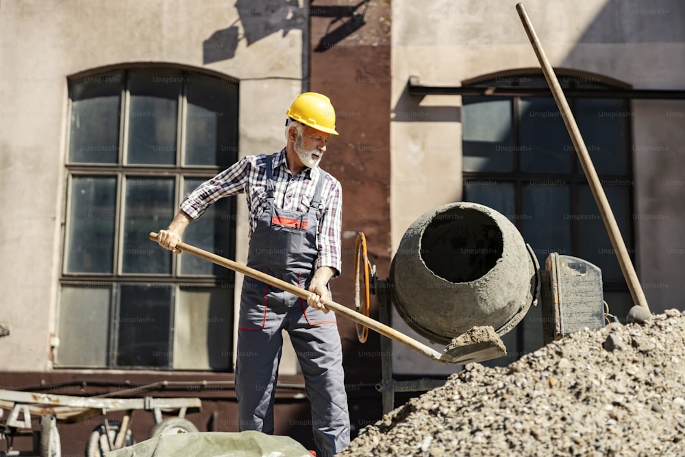 Senor Bauarbeiter vor Ort Mörtelmischer füllen. Ein leitender Bauarbeiter steht neben einem Mörtelmischer und füllt diesen auf der Baustelle mit Beton.