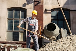 Senor construction worker on site filling mortar mixer. A senior construction worker is standing next to a mortar mixer and filling it with concrete on the construction site.