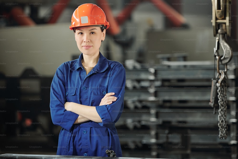 Young confident woman in hardhat, gloves and blue workwear crossing arms on chest on background of stack of details