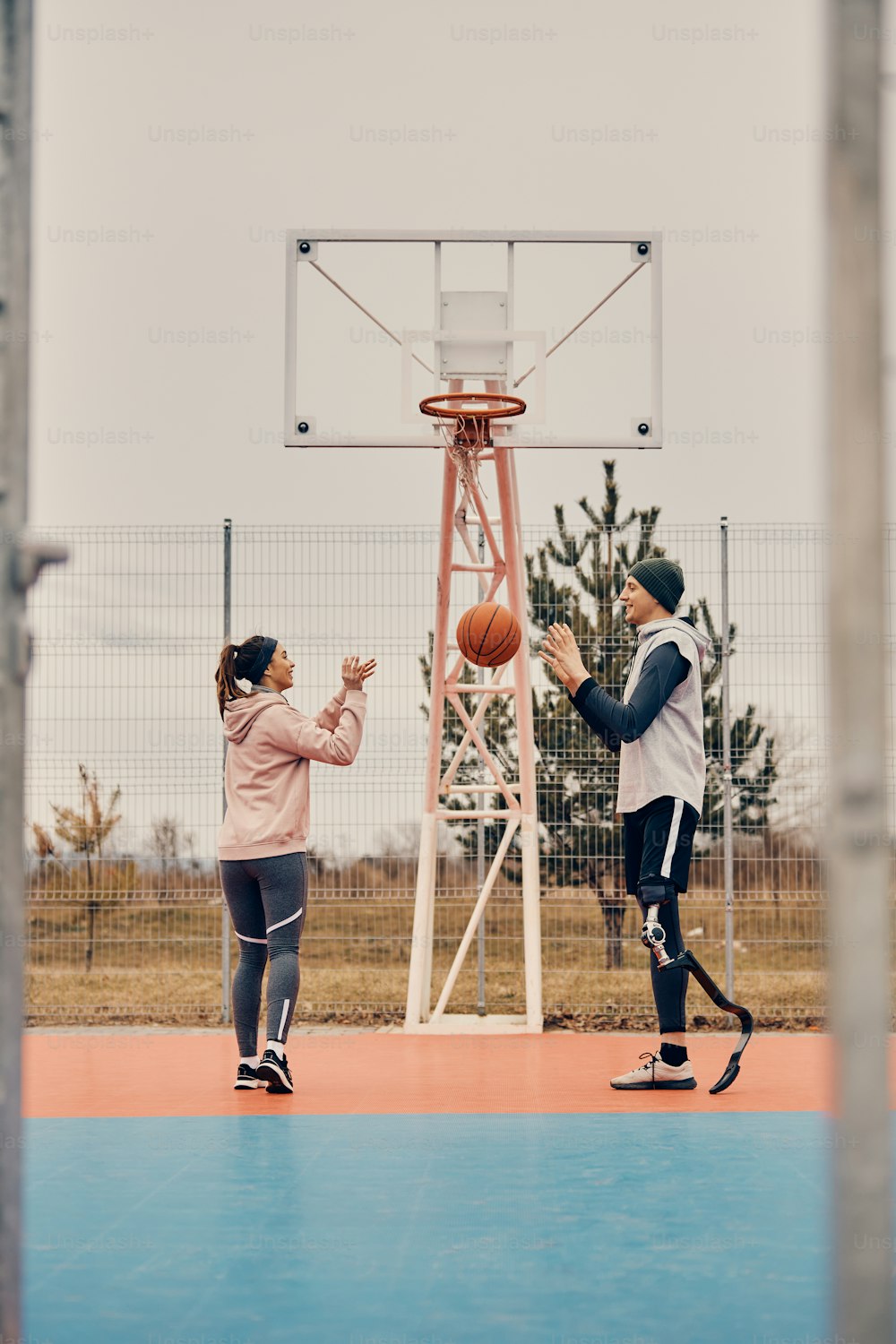 Full length of athletic woman and her male friend with leg prosthesis playing basketball on outdoors sports court.