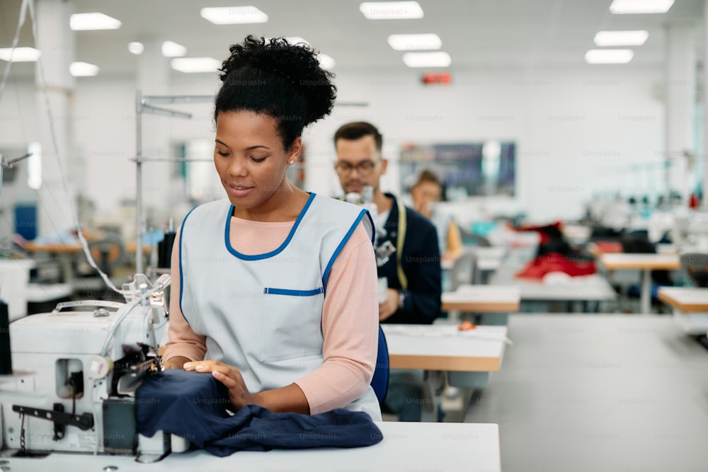 Junger afroamerikanischer Textilarbeiter näht an der Produktionslinie, während er in einer Bekleidungsfabrik arbeitet.