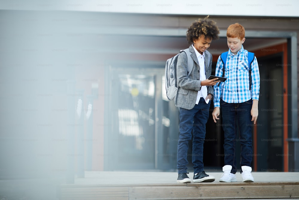 학교 아이들은 휴대 전화를 사용하여 야외에서 학교 건물 근처에서 무언가를 보면서 서 있습니다.