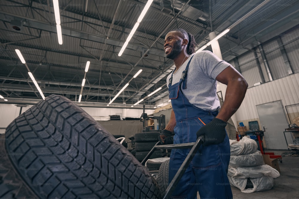 Mecânico afro-americano transportando pneus de automóveis através de oficina em carrinho com sorriso no rosto