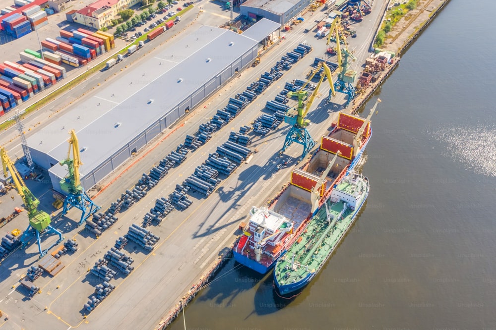 Aerial top view huge cargo ship moored at the pier at the port, loading goods, metal in aluminum rolls, concrete and other solid raw materials.