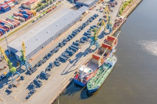 Aerial top view huge cargo ship moored at the pier at the port, loading goods, metal in aluminum rolls, concrete and other solid raw materials.