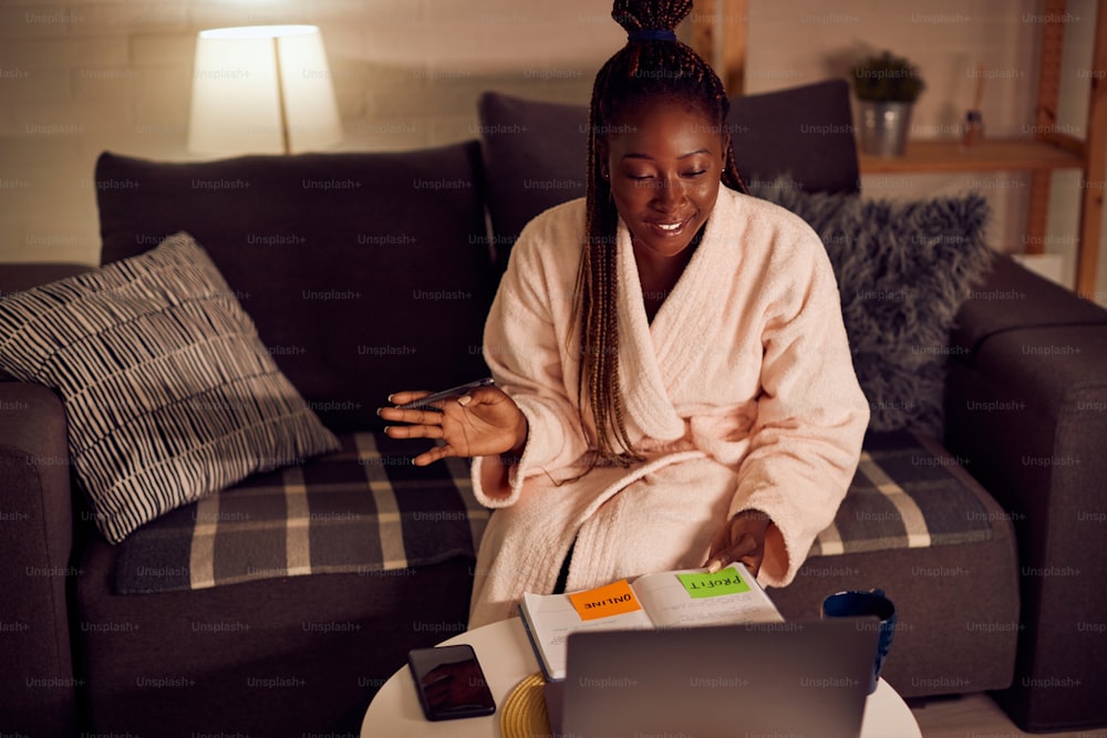 Young black woman using laptop and talking during video call while working in the evening at home.
