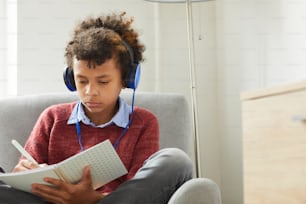 Serious African boy in headphones sitting on armchair and making notes in textbook he preparing for the lessons at home