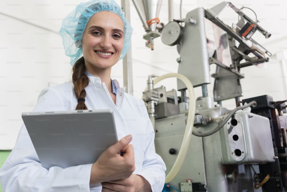 Retrato de uma inspetora de controle de qualidade sorrindo e olhando para a câmera enquanto segurava um tablet durante a inspeção em uma fábrica de cosméticos contemporânea