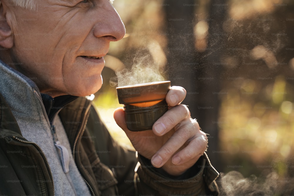 Nahaufnahme des fröhlichen älteren Mannes, der sich bei einem Spaziergang im Wald mit einer Tasse heißen Tee mit Dampf über ihr entspannt. Reisekonzept
