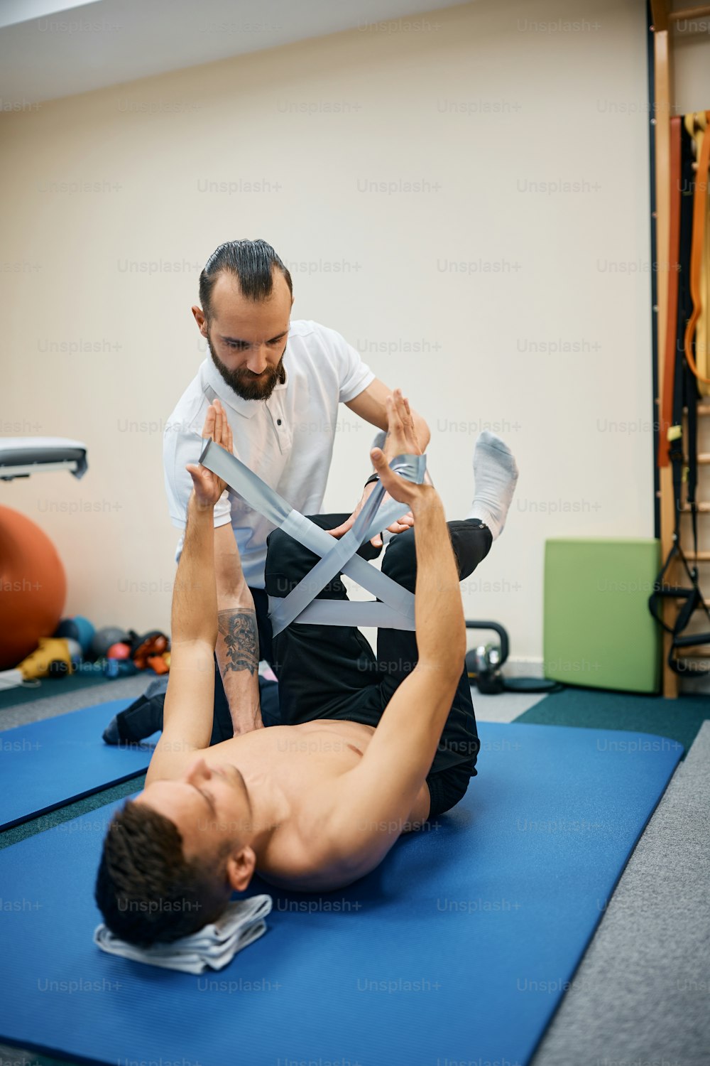 Athletic man exercising resistance band during kinesiotherapy treatment with physiotherapist at health club.