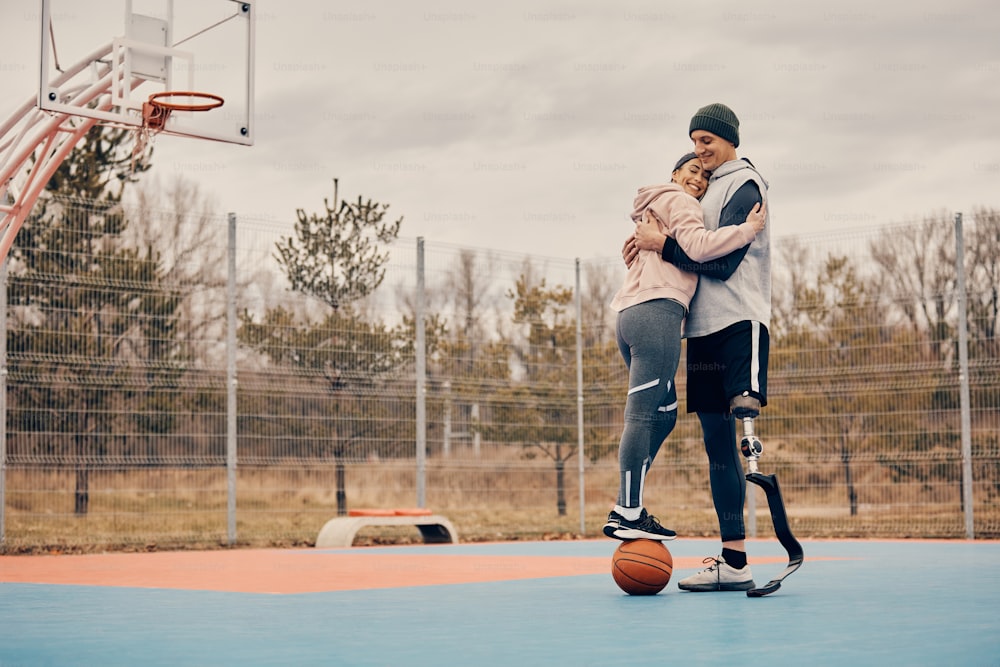 Happy athletic woman standing on a basketball while embracing with her boyfriend who has prosthetic leg on outdoors sports court.