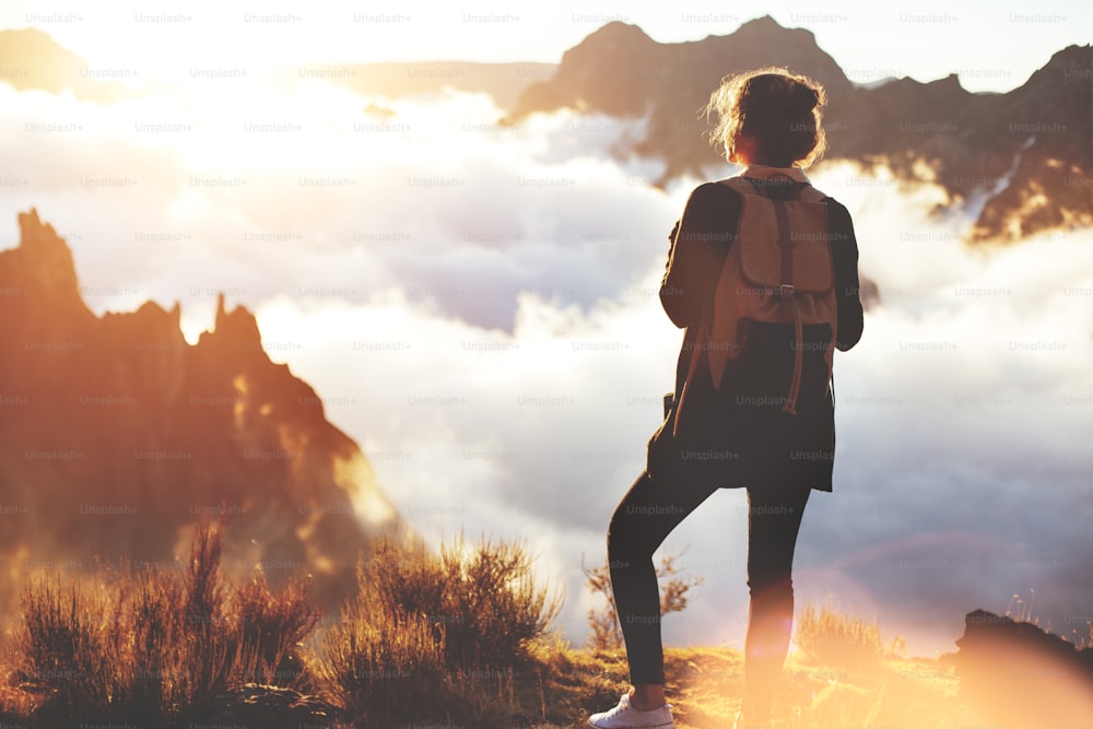 Young traveler woman with a backpack standing on edge of cliff and enjoying landscape, sunset and clouds.