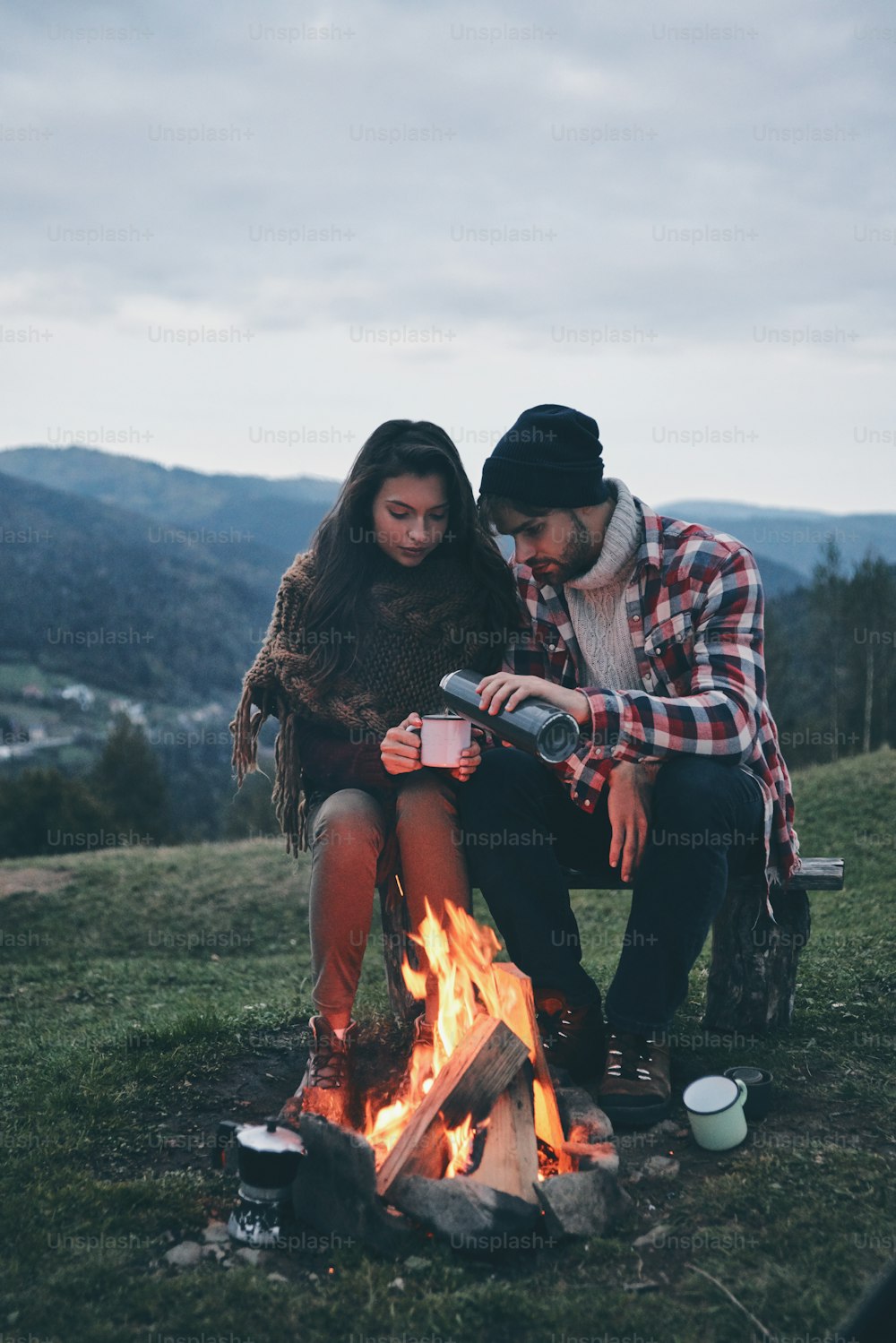 Beautiful young couple enjoying hot drinks while warming up by the campfire in the mountains