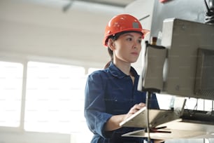 Operador asiático joven serio en casco que usa la computadora de la máquina industrial mientras elige configuraciones en el taller