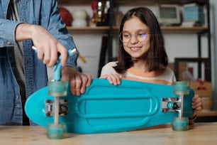 Happy youthful girl in eyeglasses looking at her father fixing wheels of blue skateboard or repairing it while standing by table in garage