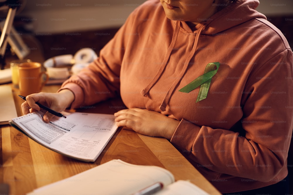 Unkenntliche Schülerin, die sich zu Hause auf bevorstehende Prüfungen vorbereitet, während sie ein grünes Band für psychische Gesundheit auf ihrem Hemd trägt.