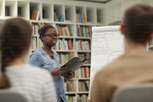 Afrikanische Smilling-Frau mit Brille liest ihre Präsentation vor jungen Geschäftsleuten in der Bibliothek