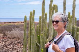 Happy senior woman with backpack enjoying outdoors excursion, cactus plant, sea and blue sky