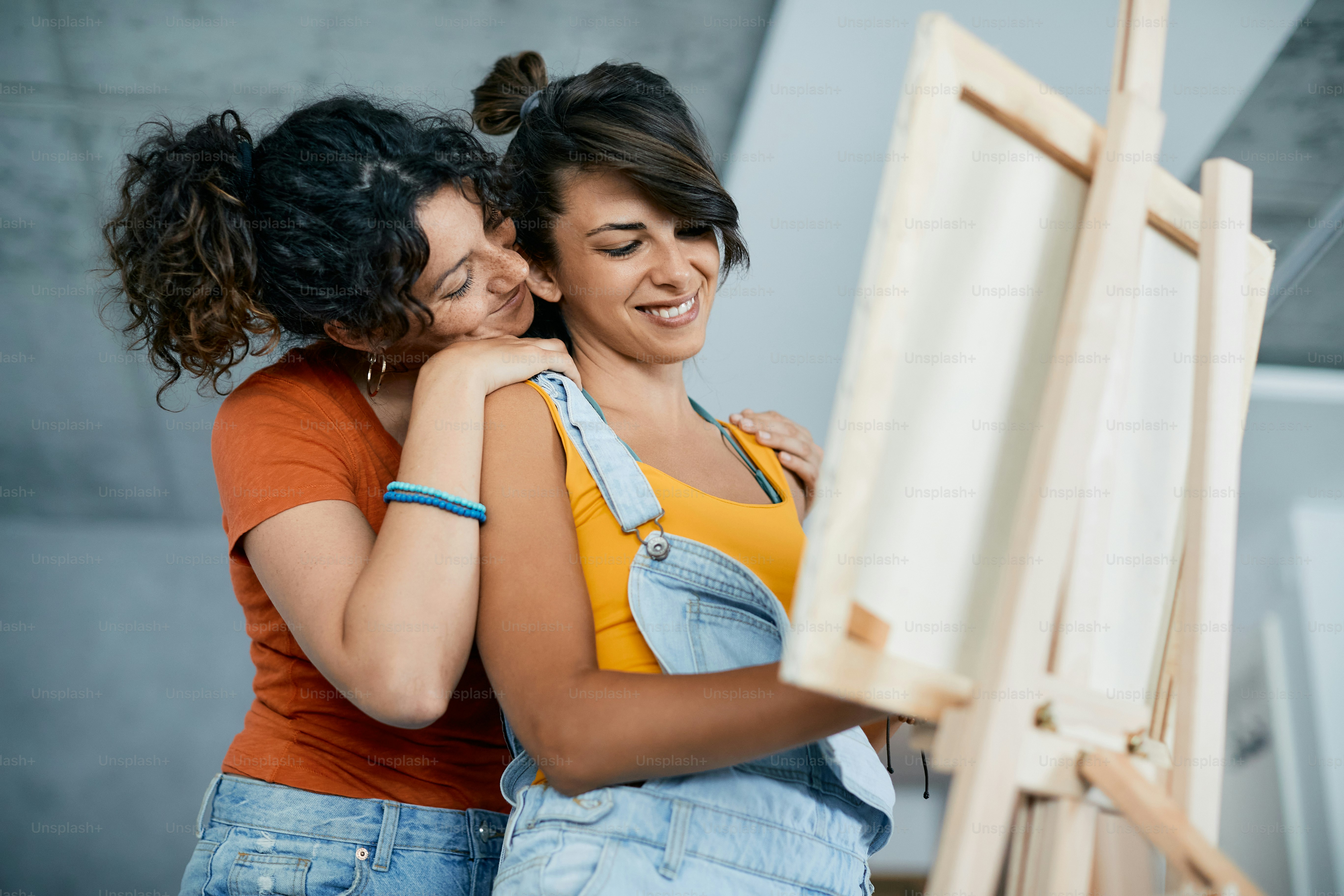 Lésbica sorridente abraça sua namorada que está pintando em tela em casa.