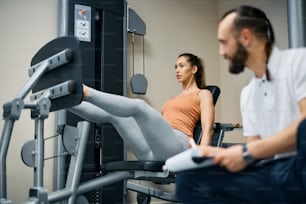 Joven atleta femenina haciendo ejercicio en la máquina de prensa de piernas mientras tiene entrenamiento deportivo con instructor de fitness en un club de salud.