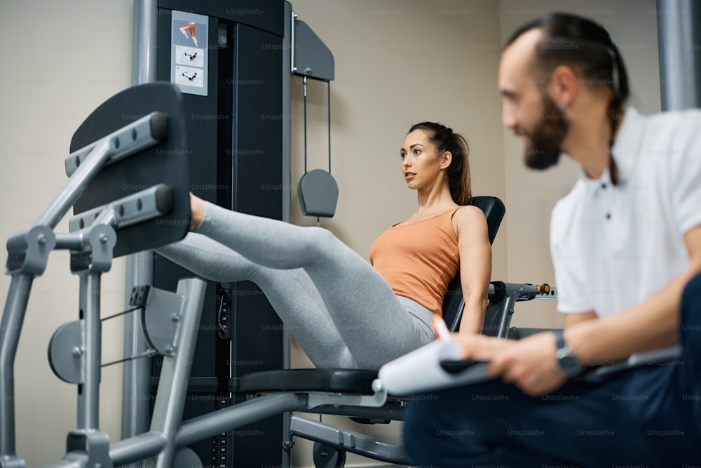 Joven atleta femenina haciendo ejercicio en la máquina de prensa de piernas mientras tiene entrenamiento deportivo con instructor de fitness en un club de salud.
