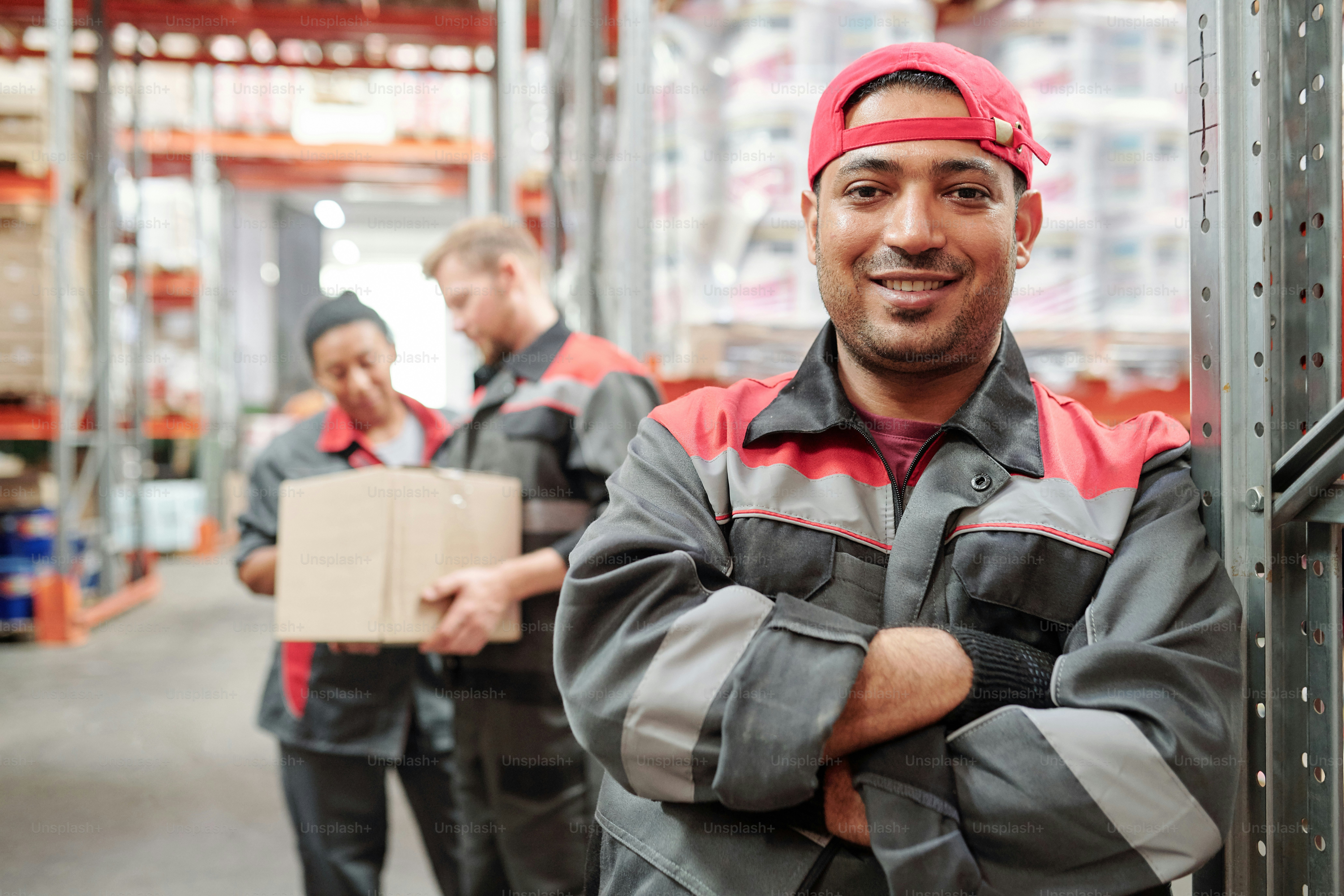 作業服と手袋を着た幸せな若いラテン系男性が、倉庫で働く同僚に立ち向かいながら、胸で腕を組んでいます