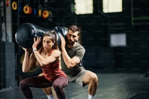 Athletic couple doing squats while holding sandbag on shoulders during gym workout. Focus is on woman.