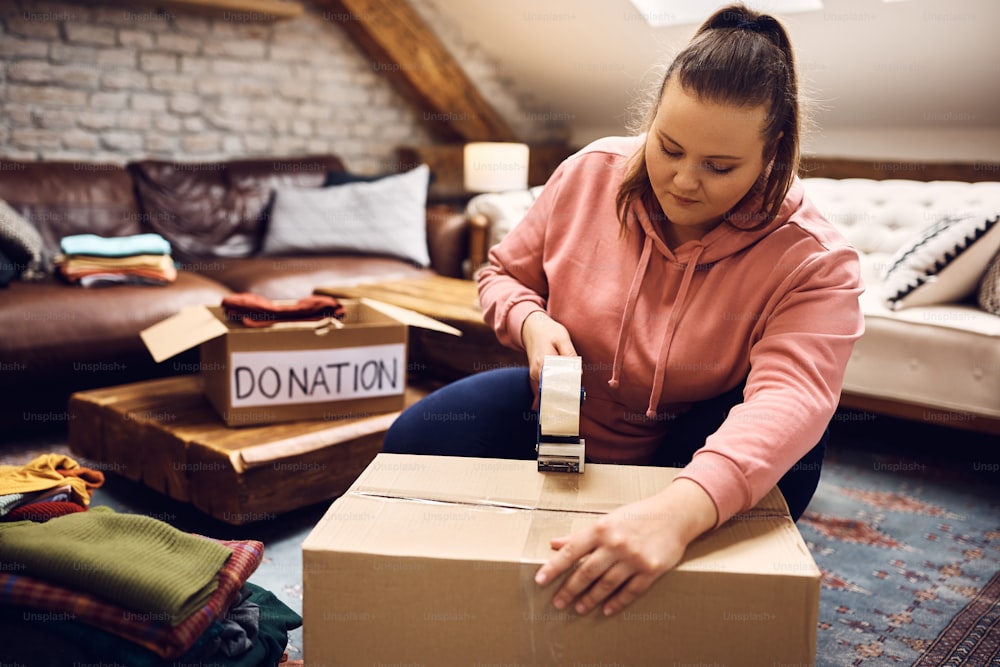 Young woman taping cardboard box with clothes to be donated to charity.