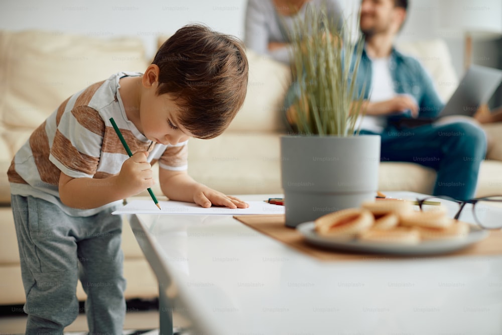 Creating little boy sketching on a piece of paper at home. His parents are in the background.