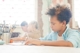 African little boy drawing at the table with other children in the background during art lesson