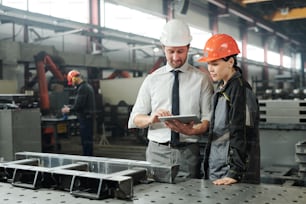 Young master in hardhat pointing at data or technical sketch on display of tablet while consulting his subordinate by workplace